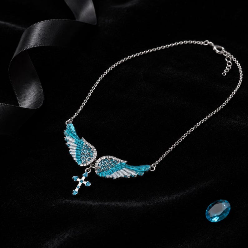 May include: A silver necklace with a pendant featuring blue and white enamel angel wings and a cross. The wings are detailed with small, sparkling stones. A loose, oval-shaped blue stone is in the lower right corner against a black background.