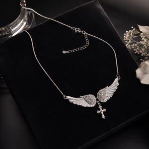 May include: A silver-toned necklace featuring a pair of detailed angel wings with a small cross pendant. The wings are embellished with small, sparkling stones. The necklace is displayed on a black surface.
