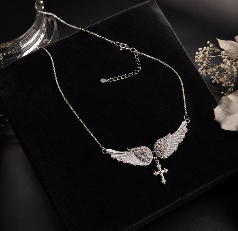 May include: A silver-toned necklace featuring a pair of detailed angel wings with a small cross pendant. The wings are embellished with small, sparkling stones. The necklace is displayed on a black surface.