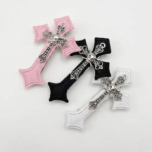 May include: Three cross-shaped hair clips in pink, black, and white. Each clip features a silver-colored skull and decorative embellishments. The crosses have a gothic aesthetic, suitable for alternative fashion.