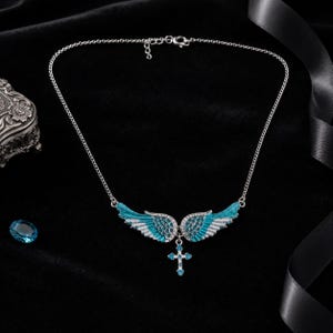 May include: A silver necklace with a pendant featuring turquoise and white wings and a cross. The wings are detailed with small, sparkling stones. The necklace is set against a black background.