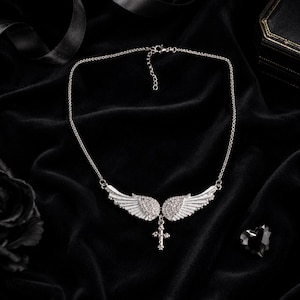 May include: A silver-toned necklace featuring a pair of detailed angel wings with a cross pendant. The wings are embellished with small, sparkling stones. The necklace is displayed on a black velvet surface.