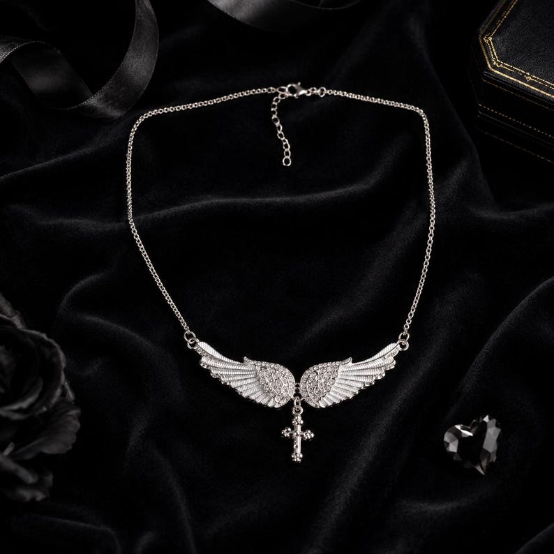 May include: A silver-toned necklace featuring a pair of detailed angel wings with a cross pendant. The wings are embellished with small, sparkling stones. The necklace is displayed on a black velvet surface.