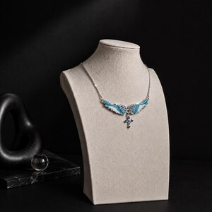 May include: A silver necklace with a pendant featuring blue and white wing-shaped designs and a cross. The necklace is displayed on a beige mannequin against a black background. The wings are detailed with small blue gemstones.