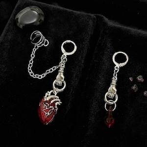May include: A pair of silver-toned earrings with a gothic theme. One earring features a red anatomical heart pendant, while the other has a red teardrop-shaped gem. Both earrings include a hand holding a ring and a chain.