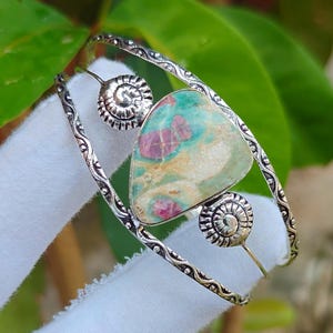 Könnte beinhalten: Ein silberfarbenes Armband in offenem Design mit komplizierten Prägemustern entlang des Bandes. Das Mittelstück ist ein dreieckiger Stein mit gesprenkelten grünen, cremefarbenen und magentafarbenen Tönen, flankiert von zwei kleinen, kreisförmigen Spiralakzenten.