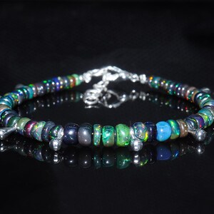 Könnte beinhalten: Ein Armband mit einer Reihe kleiner, runder, dunkel gefärbter Opalperlen mit irisierenden grünen, blauen und violetten Reflexen. Das Design enthält mehrere kleine, glatte, silberfarbene Metallanhänger in Form von Tropfen entlang des Bandes.