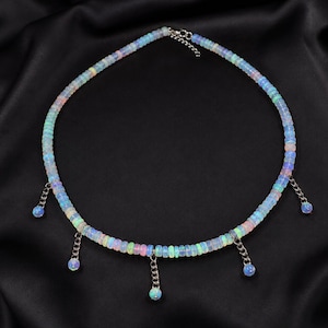 May include: A necklace featuring a strand of small, iridescent, light-colored disc beads that display flashes of blue, green, and pink. Five silver-toned metal chains hang from the strand, each ending in a single, round, iridescent bead charm.