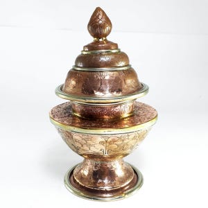 May include: A copper-colored metal lidded vessel featuring an intricate engraved floral pattern across the bowl, base, and tiered lid. The container has a wide, flared base and a pointed finial top, with polished gold-toned metal trim accents.