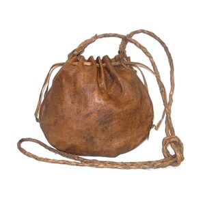 May include: A brown leather drawstring pouch with a textured surface and a gathered top closure. The bag features a long, braided leather strap that is knotted at one side, allowing for adjustable length and carrying over the shoulder.