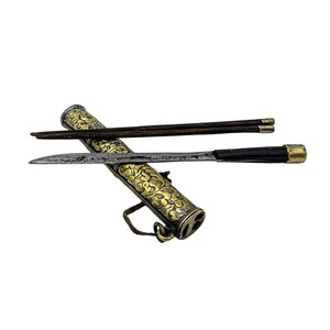 May include: A set of dark brown wooden chopsticks and a metal knife with a black handle, resting on a cylindrical metal case. The case features an intricate gold-toned floral pattern, a metallic finish, and a small loop attached to the side.
