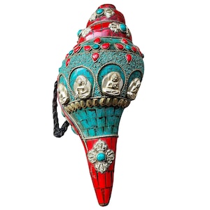 May include: A decorative conch shell, featuring a vibrant mosaic of turquoise, red, and gold accents. The shell is adorned with small turquoise and red stones, and detailed metalwork depicting seated figures. A black braided strap is attached.