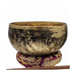 May include: A bronze-colored singing bowl with dark accents, resting on a patterned purple and gold cushion. The bowl has a wide rim and a textured surface. A wooden mallet is visible.