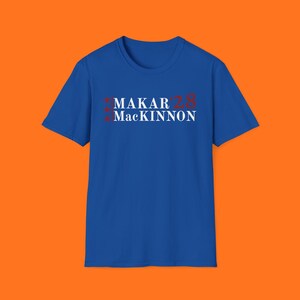 May include: A royal blue t-shirt with the name "MAKAR" and "MacKINNON" in white text. The number "28" is in red. Three red asterisks are above the name "MAKAR". The shirt is displayed on an orange background.
