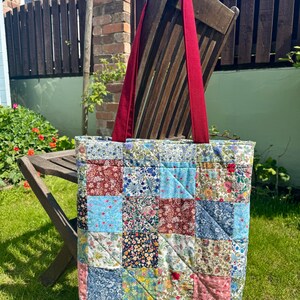 May include: A patchwork tote bag with a quilted design. The bag features a variety of floral patterns in shades of blue, pink, and red. It has a solid red strap and is hanging on a wooden chair in a garden setting.