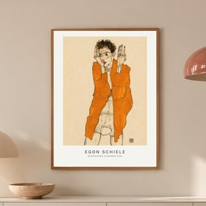 May include: A framed art print featuring a line drawing of a person wearing an orange robe, with the text EGON SCHIELE printed at the bottom. The artwork is displayed in a thin wooden frame on a light-colored wall above a white sideboard.