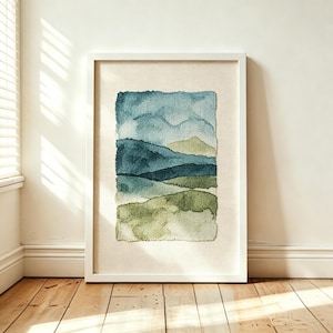 Puede incluir: Una pieza de arte mural rectangular enmarcada que presenta un bordado con aguja mágica de un paisaje. El diseño muestra colinas y montañas superpuestas en tonos verde salvia, verde bosque y azul apagado sobre una tela de color crema.