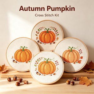 Könnte beinhalten: Vier runde Stickrahmen aus Holz mit Kreuzstich-Stoffen, die orangefarbene Kürbismotive zeigen, umgeben von verstreuten braunen Eicheln und Herbstblättern. Der Text Autumn Pumpkin Cross Stitch Kit steht oberhalb der Stickrahmen.