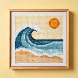 May include: A square cross-stitch embroidery in a light wood frame depicting a stylised ocean wave in shades of blue and white, a yellow sun, and a tan sandy beach against a cream background. The needlework features a clear grid pattern.