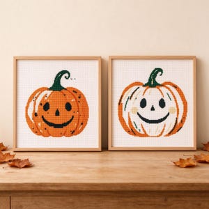 Peut inclure: Deux œuvres d'art en point de croix encadrées de forme carrée représentant des citrouilles. La pièce de gauche montre une citrouille orange avec une tige vert foncé et un visage de jack-o'-lantern noir. La droite montre une citrouille blanche.