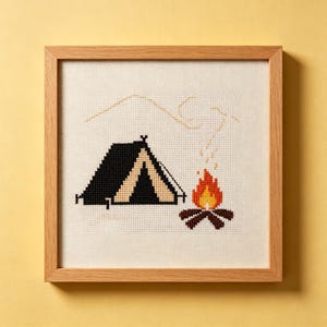 Peut inclure: Une broderie au point de croix carrée dans un cadre en bois clair représentant une tente noire avec une entrée beige, un feu de camp aux flammes orange et jaunes sur des bûches brunes, et le contour beige d'un sommet de montagne sur fond crème.