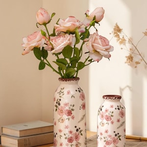May include: Two ceramic vases with a floral design. The vases are cream-coloured with pink flowers and green leaves. The rims and bases have a brown trim. The taller vase holds pink roses.