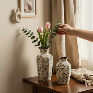 May include: Two white ceramic vases with a floral pattern, one holding pink tulips and greenery. The vases are on a wooden table, with a beige wall and curtain in the background. A small, round, white wall decoration is also visible.
