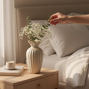 May include: A cream-colored, ribbed vase with a ruffled top holds white flowers and greenery. A small candle and a book sit on a wooden nightstand. A hand is reaching towards the flowers. The scene is set in a bedroom.