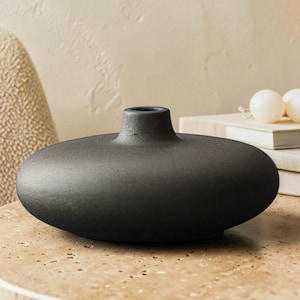 Puede incluir: Un jarrón de cerámica negro mate con un cuerpo ancho y aplanado en forma de disco y un cuello corto y estrecho. El jarrón presenta una superficie texturizada y una abertura circular en la parte superior, con una silueta de perfil bajo.