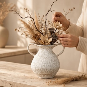 May include: A white ceramic vase with a speckled grey finish, featuring two curved handles on the sides. The vase is filled with an arrangement of dried beige flowers, pampas grass, thin brown branches, and dark brown lotus seed pods.