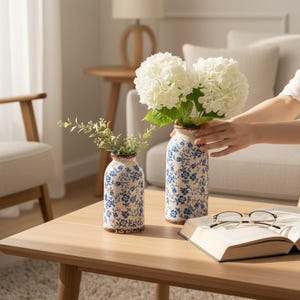 May include: Two ceramic vases with blue floral patterns and brown rims sit on a wooden table. One vase holds white flowers, the other holds greenery. A book and glasses are also on the table. The vases are a decorative home accent.