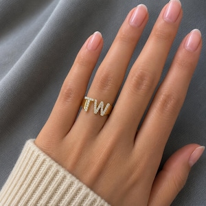Peut inclure: Une bague dorée arborant les lettres T et W comme point central. Les lettres sont ornées de petites pierres claires et scintillantes incrustées sur la surface de chaque caractère. L'anneau est lisse et poli, offrant une finition métallique nette.