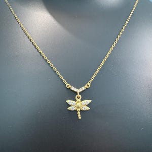 May include: A gold-toned necklace featuring a delicate chain and a dragonfly pendant. The dragonfly wings are accented with small, clear, sparkling stones. A V-shaped connector piece above the pendant is also set with tiny, clear, sparkling stones.