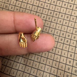 May include: A pair of gold-toned stud earrings shaped like miniature human hands. One earring depicts an open palm facing upwards, whilst the other earring shows a hand with the middle finger extended upwards, each featuring a post backing for attachment.