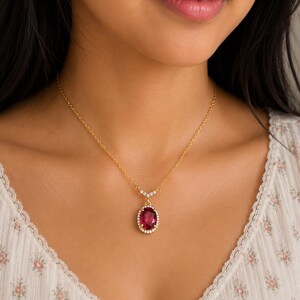 Op de afbeelding: Een gouden ketting met een ovaalvormige dieprode edelsteen hanger omringd door een halo van kleine witte steentjes. De hanger is aan de ketting bevestigd met een V-vormig oogje dat is ingelegd met piepkleine witte steentjes.