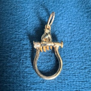 May include: A gold-toned metal pendant featuring a sculpted hand grasping a horizontal bar that connects to a U-shaped loop. The piece includes a small circular jump ring at the top for attachment, showcasing a polished metallic finish throughout.