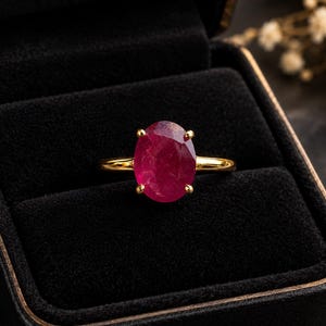 May include: A gold ring featuring a large, oval-cut, deep pinkish-red gemstone held in a four-prong setting. The band is smooth and polished gold, presented inside a black velvet-lined jewelry box.