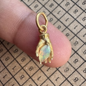 Puede incluir: Un colgante de tono dorado con forma de mano que sostiene una piedra preciosa de ópalo iridiscente de corte marquesa. El colgante cuenta con una pequeña anilla circular en la parte superior. El diseño muestra una mano detallada con dedos curvos.