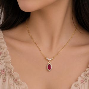 May include: A gold chain necklace featuring a marquise-cut deep red gemstone pendant surrounded by a halo of small clear stones. The pendant is attached to the chain by a small V-shaped connector adorned with three additional clear stones.