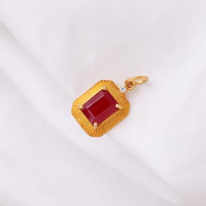 May include: A gold pendant with a rectangular, deep red gemstone at its center. The pendant has a textured, sunburst-like design around the gem and a small diamond accent. The pendant is on a white background.