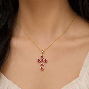 Op de afbeelding: Een gouden ketting met een kruishanger bezet met zes ronde, levendige rozerode edelstenen. Elke steen is gevat in een getextureerde gouden zetting, waardoor een kruisvorm ontstaat die hangt aan een fijne, gepolijste gouden schakelketting.