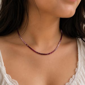 May include: A beaded necklace featuring small, round, faceted gemstones in a gradient color scheme that transitions from deep burgundy and dark red to lighter shades of pink and pale lavender, arranged in a single strand around the neck.