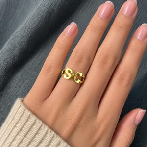 May include: A gold-toned metal ring featuring the capital letters S and C as the primary design element. The letters are connected to form a continuous band that wraps around the finger, showcasing a polished, reflective surface finish.