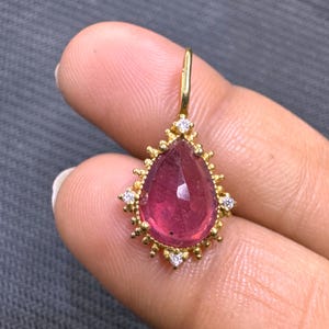 Pode incluir: Pingente de ouro em forma de gota, com uma gema rosa vibrante. A moldura dourada é adornada com pequenos diamantes brilhantes. O pingente é suspenso por uma argola de ouro.