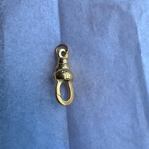 Pode incluir: Um fecho lagosta giratório de metal dourado com uma secção superior arredondada e escalonada e um mecanismo de mola. A peça tem uma superfície polida e refletora, incluindo um pequeno anel circular preso à argola superior.