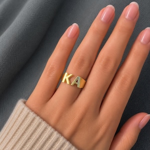 Op de afbeelding: Een goudkleurige ring met de hoofdletters K en A als centraal designelement. De letters zijn met elkaar verbonden en liggen horizontaal op de vinger, waarbij het oppervlak van de ring een gladde, gepolijste metalen afwerking vertoont.