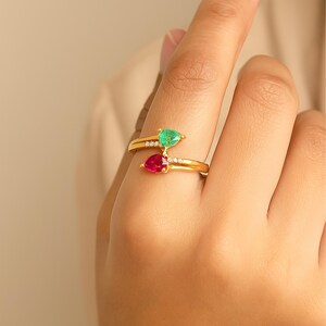 Puede incluir: Un anillo de oro tipo bypass con una piedra preciosa verde en forma de pera y una piedra preciosa roja en forma de pera, cada una acentuada por pequeñas piedras claras a lo largo de la banda. El anillo tiene un acabado dorado pulido y abierto.