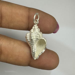 Può includere: Un ciondolo a forma di conchiglia color argento con una superficie strutturata e un piccolo anello nella parte superiore. Il ciondolo è tenuto tra le dita, mostrando i suoi dettagli intricati e la sua forma naturale. La conchiglia è di colore argento chiaro.