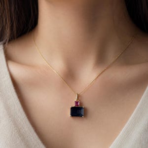 May include: A gold necklace with a pendant featuring a large, rectangular, dark blue gemstone below a smaller, rectangular, pink gemstone. The necklace is worn against a light-colored top.