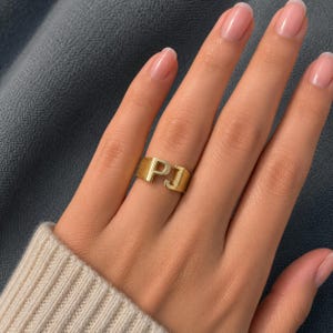 Peut inclure: Une bague dorée arborant les lettres majuscules P et J comme élément central. L'anneau présente une finition lisse et polie sur les lettres et une surface texturée et mate sur les côtés, créant un contraste marqué dans la réflexion de la lumière.
